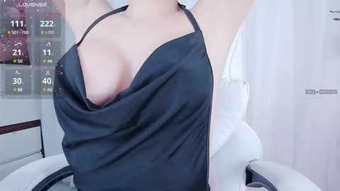 Media: A video of a woman with fair skin, wearing a black, low-cut apron dress, revealing ample cleavage. Background shows a white room with a digital display and a white chair.