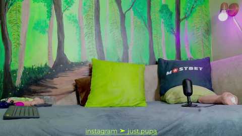 Media: Video of a cozy bedroom with a green forest mural, green and yellow pillows, a beige blanket, and a black microphone on a grey bed.