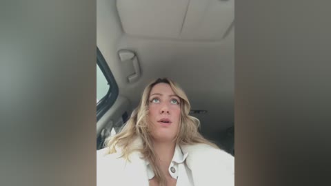 Media: Video of a surprised blonde woman with long wavy hair, wearing a white shirt, sitting in a car with beige interior.