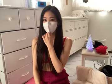 Media: Video of an Asian woman with long black hair, wearing a red dress and face mask, sitting in a brightly lit room with white furniture, a small table, and a purple lamp.