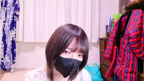 Media: Video of an East Asian woman with straight, shoulder-length brown hair, wearing a black face mask, white lace top, and standing in a bedroom with hanging clothes and a floral curtain.