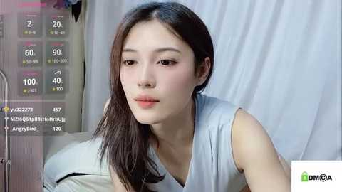 Media: Video of a young East Asian woman with long dark hair, wearing a sleeveless light gray top, sitting on a bed. Background includes a digital display with health metrics and a white curtain.