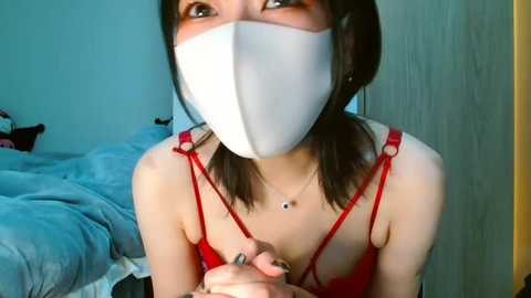 Media: Video of an Asian woman with a white mask, red lingerie, and a necklace, kneeling in a dimly lit bedroom with a blue bed and wooden door.