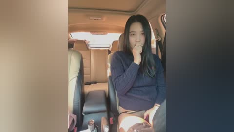 Media: Video of a young Asian woman with long black hair, wearing a dark sweater, sitting in a car seat, holding her face, looking thoughtful.