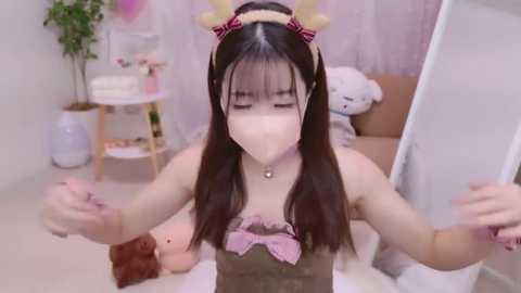 Media: Video of a young Asian woman with long black hair, wearing a pink bow headband, a mask, and a pink lace top, posing with arms outstretched in a pastel bedroom with soft, pink and white decor.