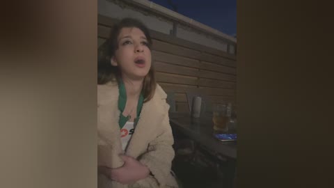 Media: A video of a woman, possibly in her 30s, with long brown hair, wearing a white robe, and a green shirt, looking surprised and holding her stomach, in a dimly-lit outdoor setting with a wooden fence and beer bottles on a table in the background.