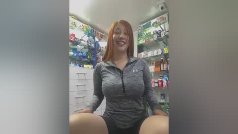 Media: Video of a smiling, red-haired woman in a grey athletic top and black shorts, sitting in a brightly lit pharmacy with shelves full of various products.