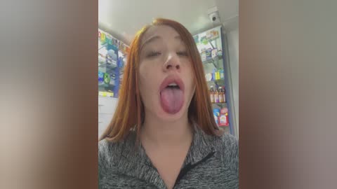 Media: Video of a young Asian woman with fair skin and straight, reddish-brown hair, sticking her tongue out and wearing a gray polo shirt. Background shows a brightly lit, cluttered room with colorful toys and shelves.