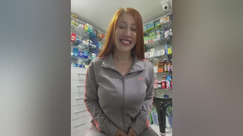 Video of a smiling, fair-skinned, red-haired woman in a gray zip-up jacket, standing in a brightly lit pharmacy with shelves full of colorful medication.