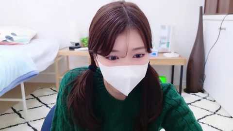 Media: A video of a young Asian woman with long brown pigtails, wearing a green sweater and a white face mask, seated indoors in a minimalist bedroom with a bed, nightstand, and white patterned rug.