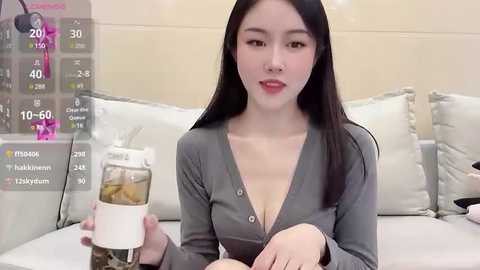 Media: Video of an East Asian woman with long black hair, light skin, and red lipstick, wearing a grey cardigan, sitting on a white couch, holding a glass of iced tea.