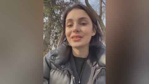 Media: Video of a young woman with fair skin, dark hair, and light makeup, wearing a black puffer jacket, smiling outdoors amidst bare trees and green foliage.