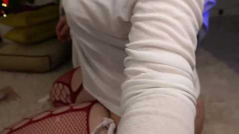 Media: Video of a person with fair skin and a white long-sleeve shirt, kneeling on a beige carpet, wearing red fishnet stockings, in a dimly lit room with a green couch and festive lights.