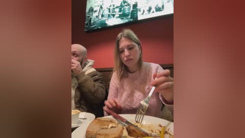 Media: Video of a woman with blonde hair and a pink sweater eating a large pancake with a fork at a diner. Behind her, a bald man in a brown jacket and another person are seated.