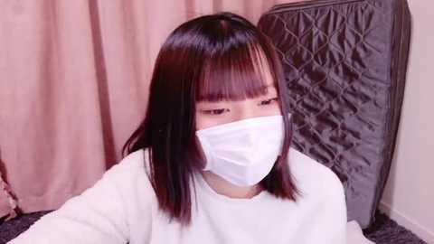 Media: Video of an East Asian woman with straight, shoulder-length black hair, wearing a white face mask, white top, and gray quilted blanket, indoors with light pink curtains and carpeted floor.