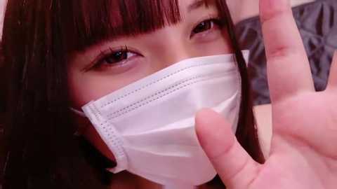 Media: A close-up video of an Asian woman with straight, dark hair and blunt bangs, wearing a white surgical mask, with a man's hand pointing at her face.