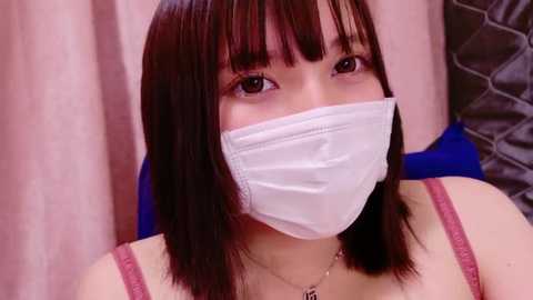 Media: Video of an Asian woman with straight black hair, wearing a white surgical mask, pink spaghetti strap top, and a gold necklace. She has fair skin and sits in front of a pink and blue patterned background.