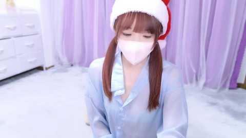 Media: Video of a young Asian woman with fair skin, wearing a Santa hat, white mask, and see-through blue blouse. Background features white drawers and lavender curtains in a brightly lit room.