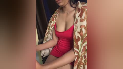 Media: Video of a woman with medium skin tone, wearing a red mini dress and a patterned shawl, sitting on a chair with a blurred background.