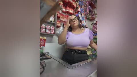 Media: Video of a Latina woman with medium skin tone, wearing a lavender crop top and black pants, standing behind a candy store counter. She has curly dark hair and is adjusting her glasses.