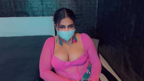 Media: Video of a South Asian woman with medium skin tone, wearing a pink lace bralette, teal mask, and long earrings, sitting on a bed with a dark wall in the background.