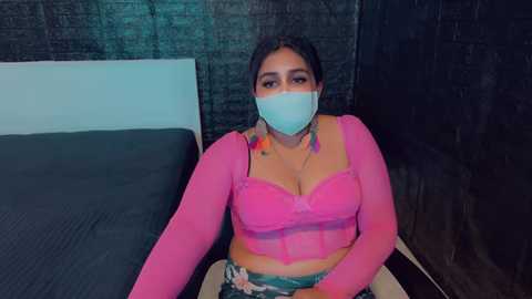 Media: A video of a curvy, dark-skinned woman in a pink lace bra and matching mask, seated on a bed with a teal pillow and dark bedspread.