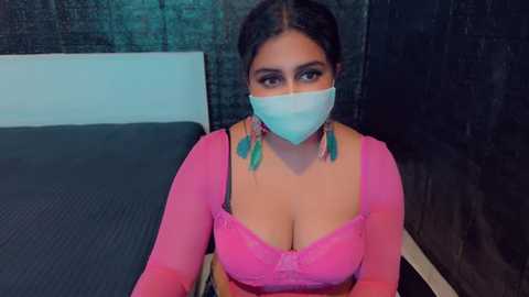 Media: A video of a South Asian woman with dark hair and medium skin tone, wearing a pink bra, teal earrings, and a blue mask, sitting on a bed with a dark headboard.