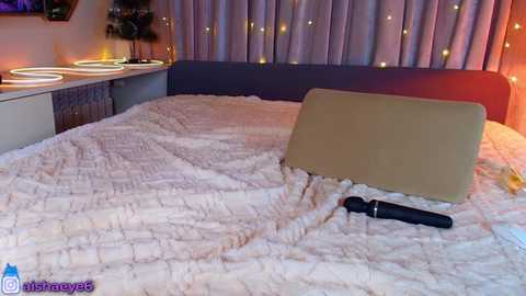 Media: Video of a modern bedroom with a beige quilted bedspread, a tan pillow, a black vibrator on the bed, and a cozy ambiance with string lights and a TV in the background.