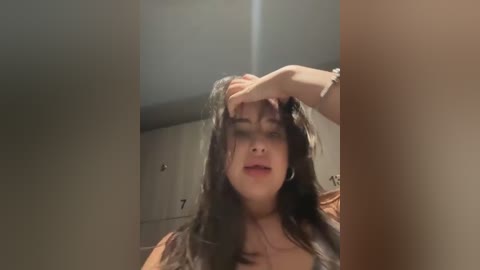 Media: Video of a young woman with long dark hair, light skin, and a medium build, wearing a brown top. She appears distressed, with one hand on her head, in a dimly lit room with beige walls.