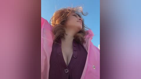 Media: Video of a young woman with light brown hair, wearing a pink raincoat over a dark purple knit sweater, against a clear blue sky.