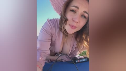 Media: Video of a young woman with fair skin and wavy brown hair, wearing a pink hat and shirt, leaning over a blue table. Background includes a clear blue sky and green grass.