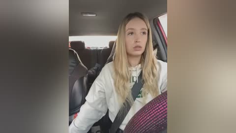 Media: Video of a young Caucasian woman with long, blonde hair, wearing a white sweater, seated in a car with a black seatbelt. The background shows the car's interior with dark upholstery and a blurred, beige wall.