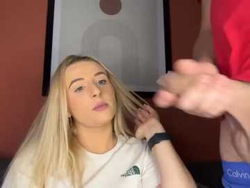 Media: Video of a blonde woman with fair skin, wearing a white t-shirt, sitting in a salon chair, with a hand brushing her hair. A man in red clothing stands in the background.