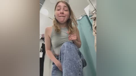 Media: Video of a young woman with long blonde hair, wearing a grey crop top and high-waisted jeans, posing in a small, brightly lit bathroom with teal walls and a white towel.