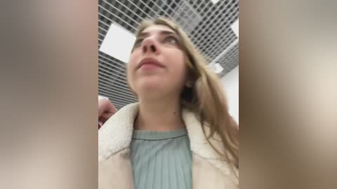 Media: Video of a young woman with long, light brown hair, wearing a light blue shirt and a beige coat, looking upward in a modern office with a grid-patterned ceiling.