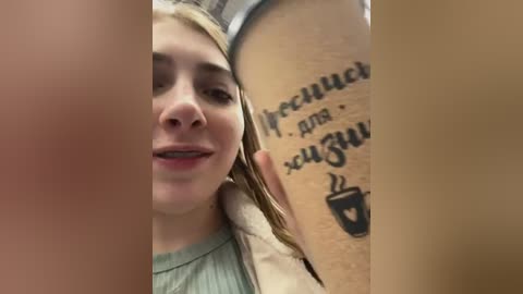 Media: Video of a young woman with light skin and long blonde hair, smiling while holding a coffee cup with a tattoo on her arm.