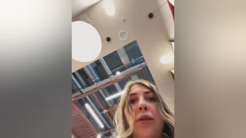 Media: Video of a young woman with long blonde hair, wearing a light jacket, standing in a modern, well-lit room with white walls, round ceiling lights, and tall bookshelves.