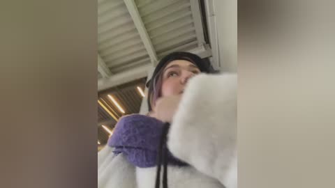 Media: Video of a young person with fair skin, wearing a purple beanie and white hoodie, looking up in a brightly lit room with horizontal blinds.