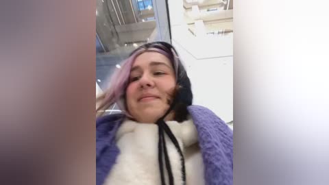 Media: Video of a person wearing a purple, fuzzy, hooded costume with a black hood and white inner garment, sitting in a modern, glass-walled building. The person has light skin and a slight smile.