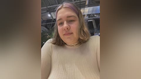 Media: Video of a young Caucasian woman with shoulder-length blonde hair, wearing a beige ribbed sweater, standing in a modern industrial setting with metal beams and a green plant in the background.