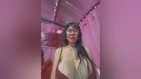 Media: Video of a woman with long black hair, wearing glasses and a low-cut red dress, standing in a pink-themed tent with white string lights.