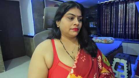 Media: Video of a middle-aged Indian woman with long black hair, wearing a red sari with floral patterns, sitting in a dimly lit room with brown curtains and a white bed.