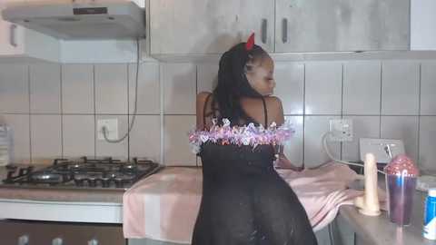 Media: Video of a Black woman with long braids, wearing a black dress with floral print, sitting on a kitchen counter in a white-tiled kitchen.