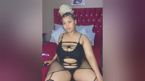 Media: A video of a plus-size, dark-skinned woman with braided hair, wearing black lingerie with fishnet stockings, sitting on a bed with red bedding.