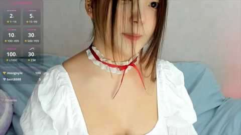 Media: Video of a young Asian woman with light skin and brown hair, wearing a white blouse with red ribbon choker, sitting on a bed with blue sheets, in a bedroom setting.