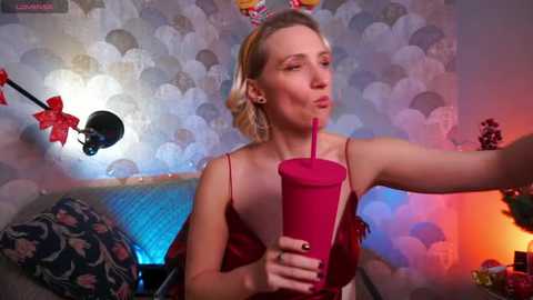 Video of a blonde woman with a slender physique, wearing a red satin robe, holding a red plastic cup, in a room with patterned wallpaper, red balloons, and a blue light.