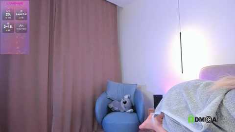 Media: A video of a cozy bedroom with a light blue armchair, beige curtains, a white wall, and a hanging lamp. A small grey dog is sleeping on the chair.