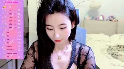 Media: Video of a young Asian woman with long black hair, wearing a sheer black lace top, sitting in a bedroom with a messy bed, stuffed animals, and a blue chair.