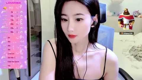 Media: Video of an East Asian woman with long black hair, fair skin, and red lipstick, wearing a black spaghetti strap top, sitting indoors. Background includes a pink calendar, a white bed, and a video game character figurine.