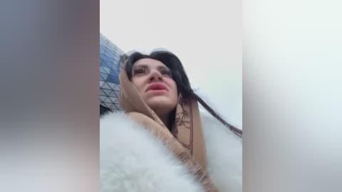 Media: A close-up video of a woman with long, dark hair, wearing a brown top and a fluffy white blanket. She looks up, eyes half-closed, in a slightly blurred, intimate setting.